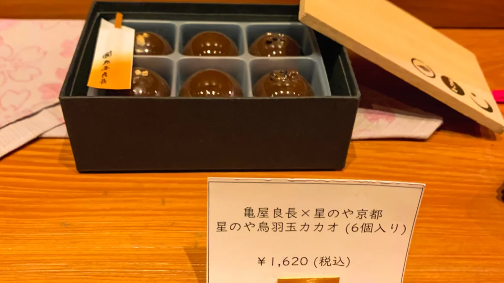 An exclusive collaboration sweets box from Kameya Yoshinaga and Hoshinoya Kyoto containing six glossy dome-shaped chocolate bonbons in a dark box with a wooden lid.

