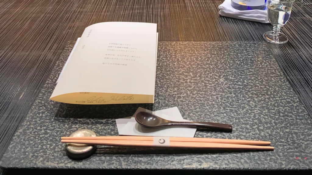 A formal kaiseki place setting with a slate mat, a menu card, and a crescent-moon shaped chopstick rest.