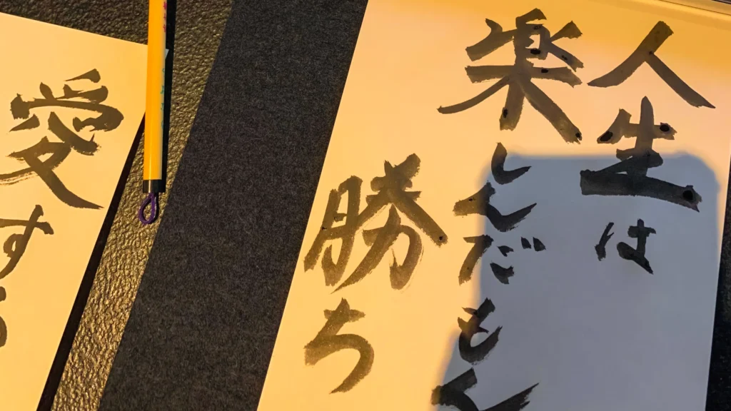 A completed piece of Japanese calligraphy reading '人生は楽しんだもん勝ち' (The one who enjoys life wins).