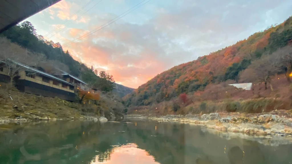 View through the boat window showing the traditional wooden buildings of Hoshinoya Kyoto among autumn trees above the crystal clear river.


