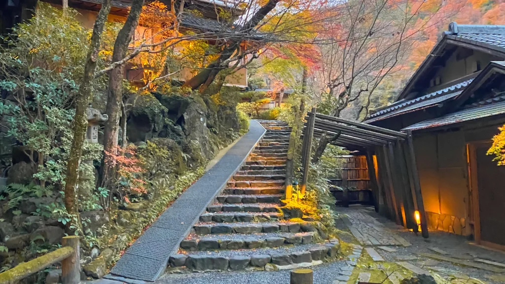 A stone stairway lined with mossy rocks, stone lanterns, and autumn foliage glowing in amber light.