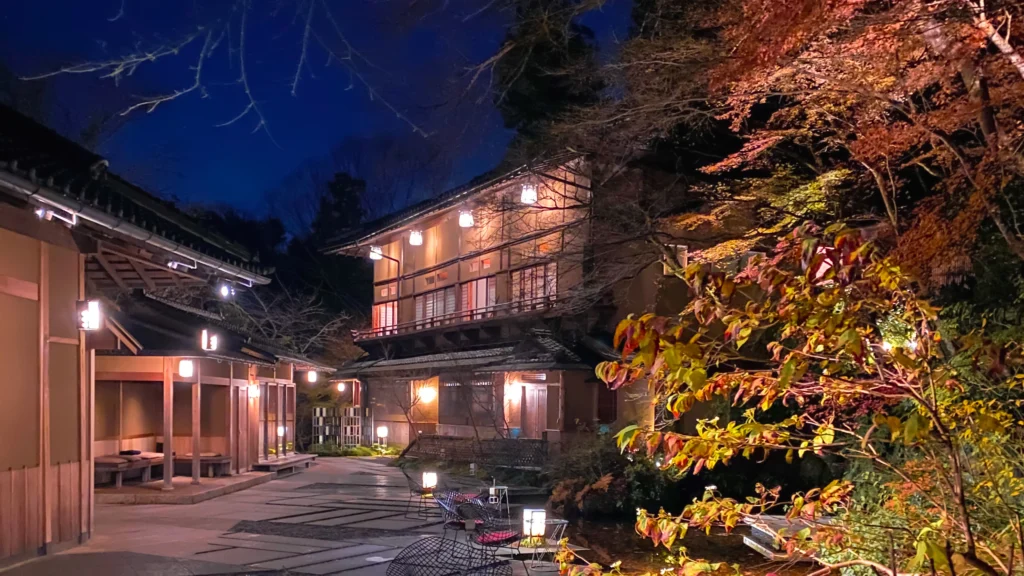 The exterior of a lantern-lit wooden building at night surrounded by autumn foliage and twilight skies.
