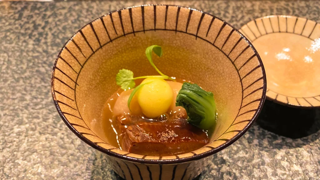 The Nimono simmered dish featuring braised meat and a yuzu-an ball in a striped ceramic bowl.