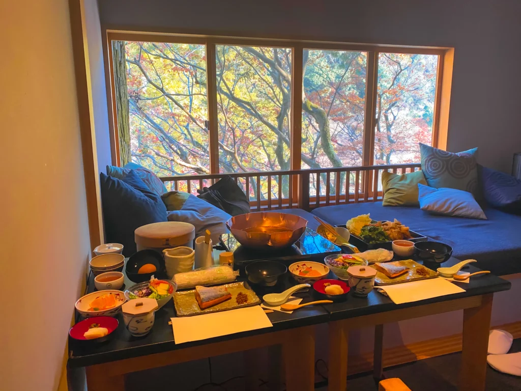 A lavish Japanese breakfast setup in the room featuring a copper nabe hot pot on an IH burner, numerous small dishes, and a view of autumn foliage through the windows.