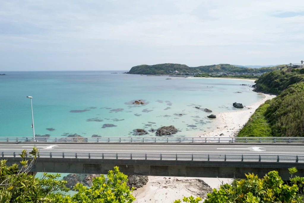 View looking down from above the bridge at the coastline, showing turquoise shallow water with scattered dark rocks, a curved beach, the bridge railing in the foreground, and the far coast of the mainland visible in the distance