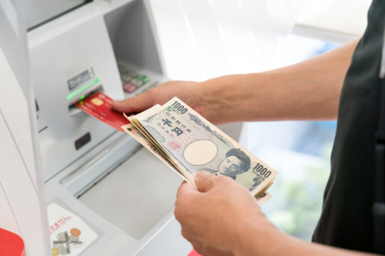 Hands inserting a red bank card into a Japanese ATM while holding a stack of 1000-yen banknotes