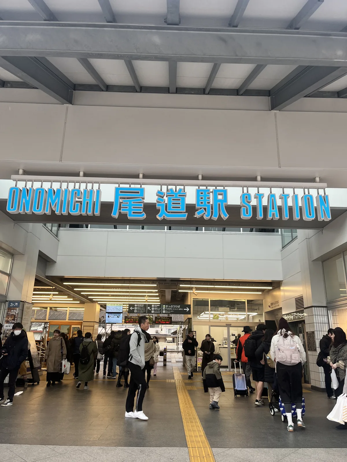The Onomichi Station entrance with passengers entering and a large blue sign reading 'ONOMICHI 尾道駅 STATION'