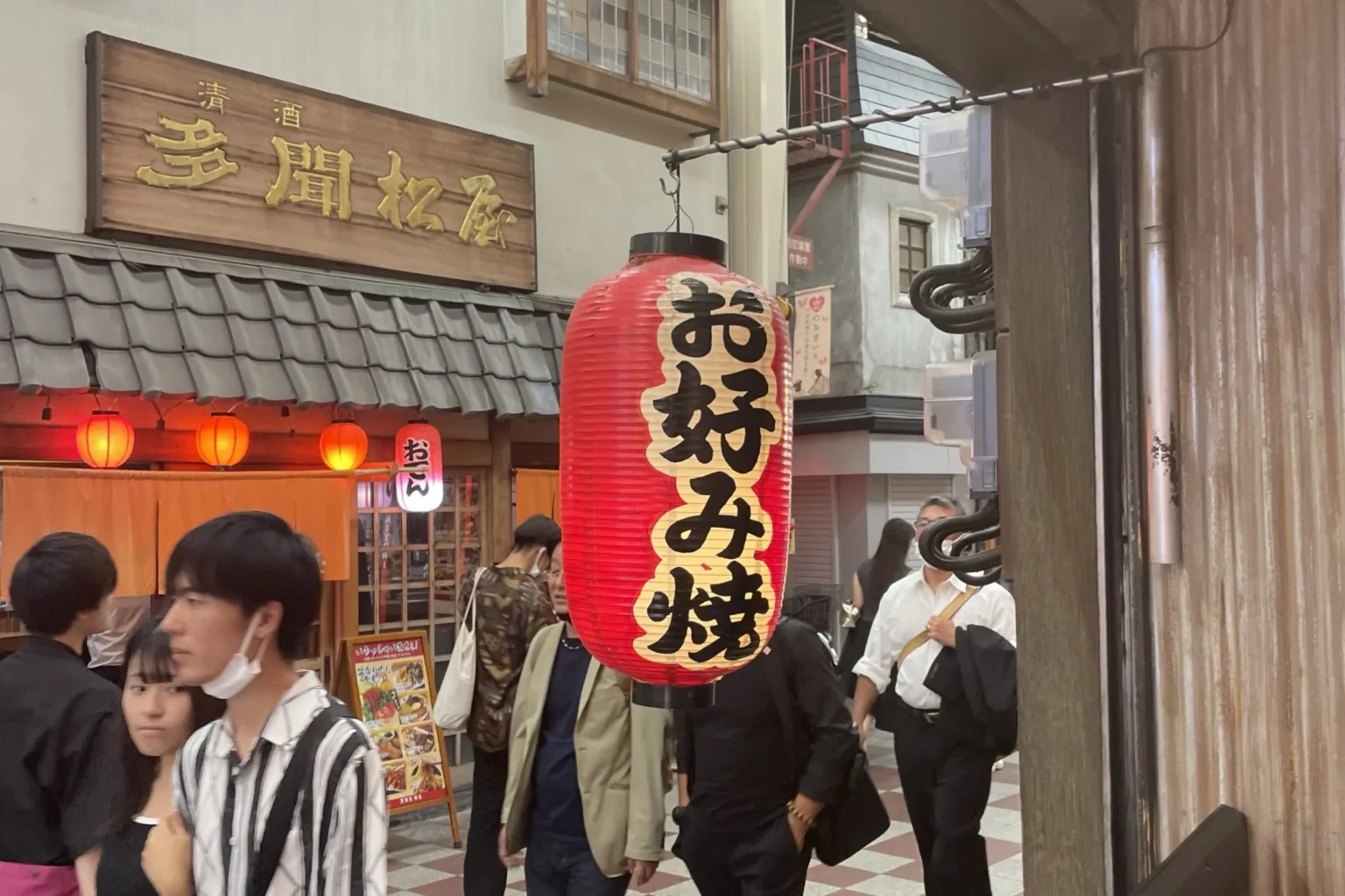 A red okonomiyaki paper lantern glowing in a narrow Dotonbori side alley at dusk in Osaka with restaurant signs stacked behind it