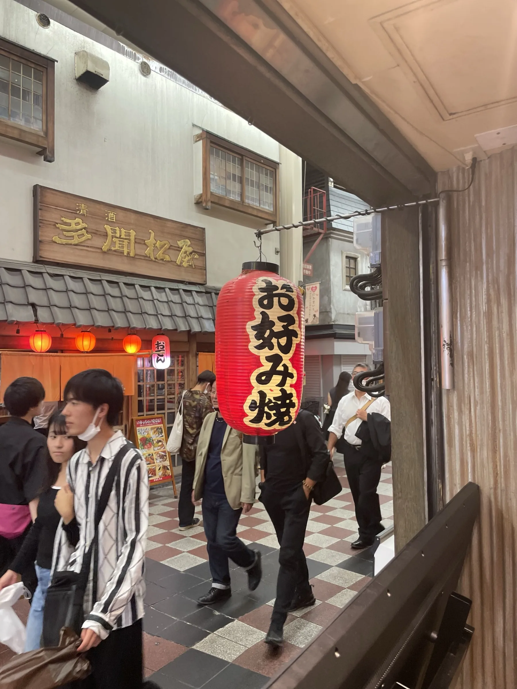 Tall red okonomiyaki paper lantern hanging in a Dotonbori side-alley with salarymen walking past