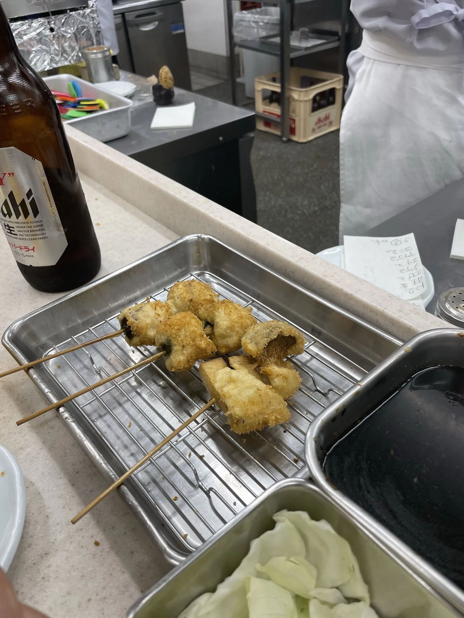Just-fried kushikatsu skewers on a stainless rack with Asahi beer bottle and cabbage on an Osaka counter