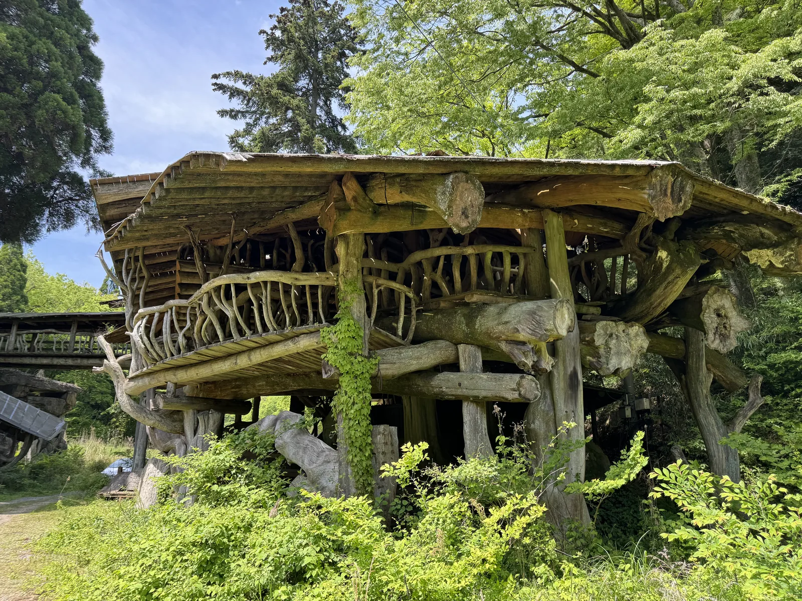 The Takamori Tree House structure photographed from the side showing the wooden balcony rails layered above the rocky stone base and surrounded by spring greenery