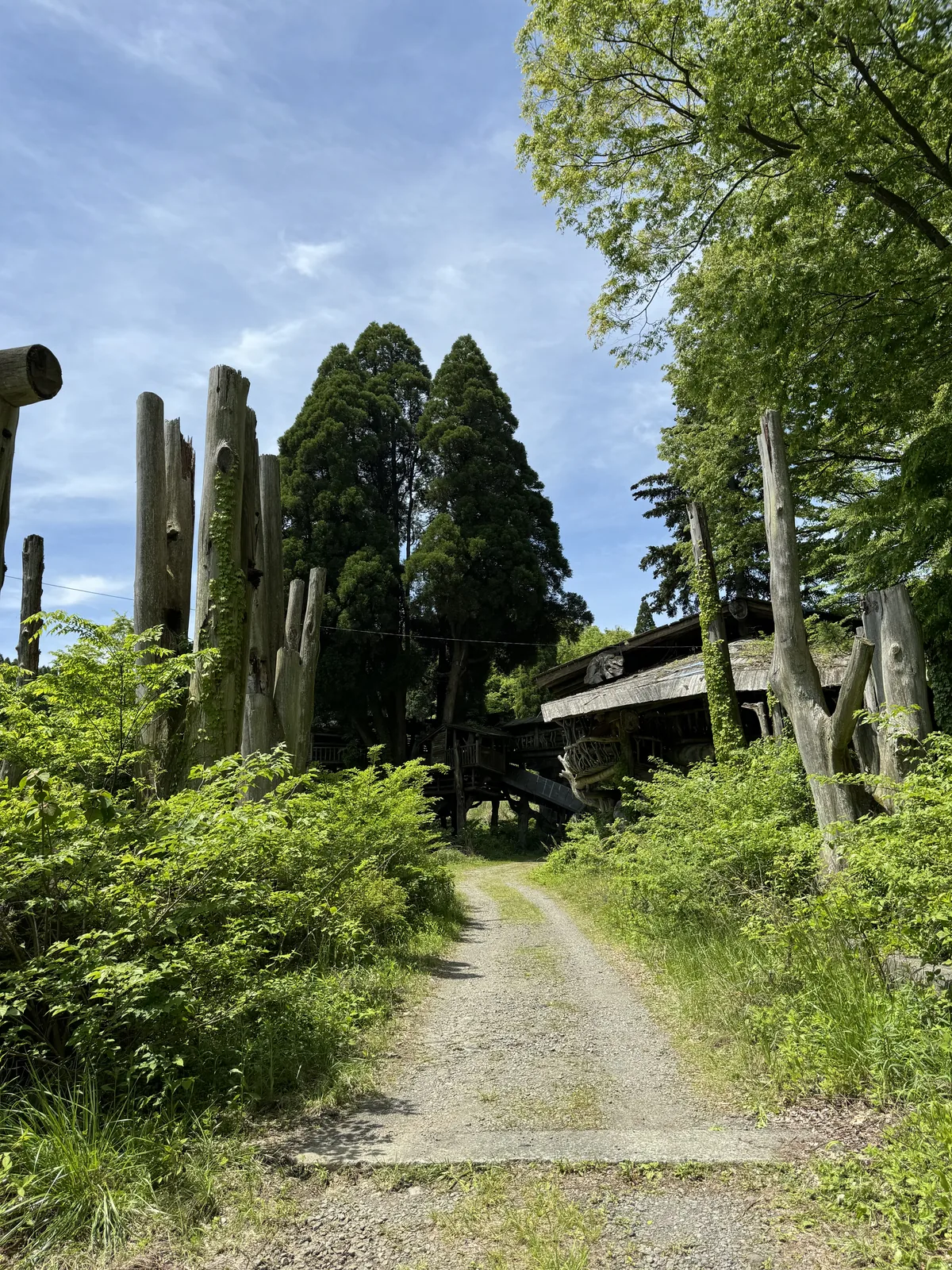 The wooden post entrance to the Takamori Tree House grounds with vertical timber pillars marking the gravel approach path through fresh spring greenery