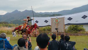 Takeda-style yabusame archer at full draw on a brown horse with red drape, target curtain to the right with concentric tomoe pattern, mountains and lake in the background, audience silhouette in foreground