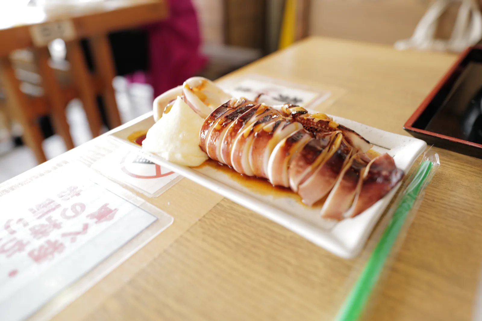 Whole grilled squid (maru-yaki ika) sliced into rings on a white plate with grated daikon radish and soy-sauce glaze, served on a wooden counter at a Nakamise-dori shop in Enoshima