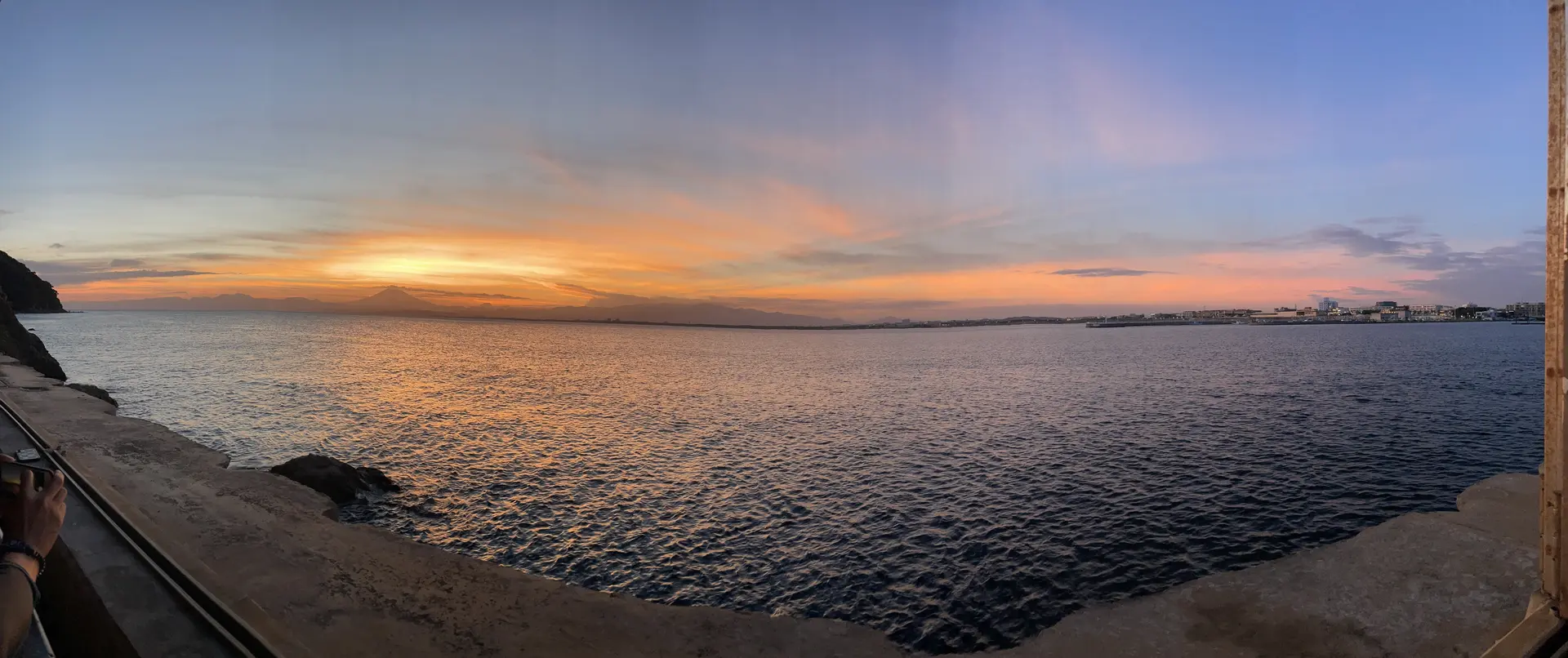 Panoramic sunset showing the curve of Sagami Bay from the Enoshima Island Spa seawall, with Mt. Fuji silhouette in the distance and Hayama and Zushi city lights visible on the right shoreline