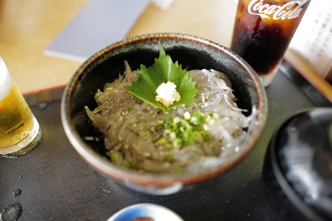 A bowl of fresh raw shirasu (whitebait) over rice topped with shiso leaf, grated ginger, and chopped scallion, served alongside iced cola and beer
