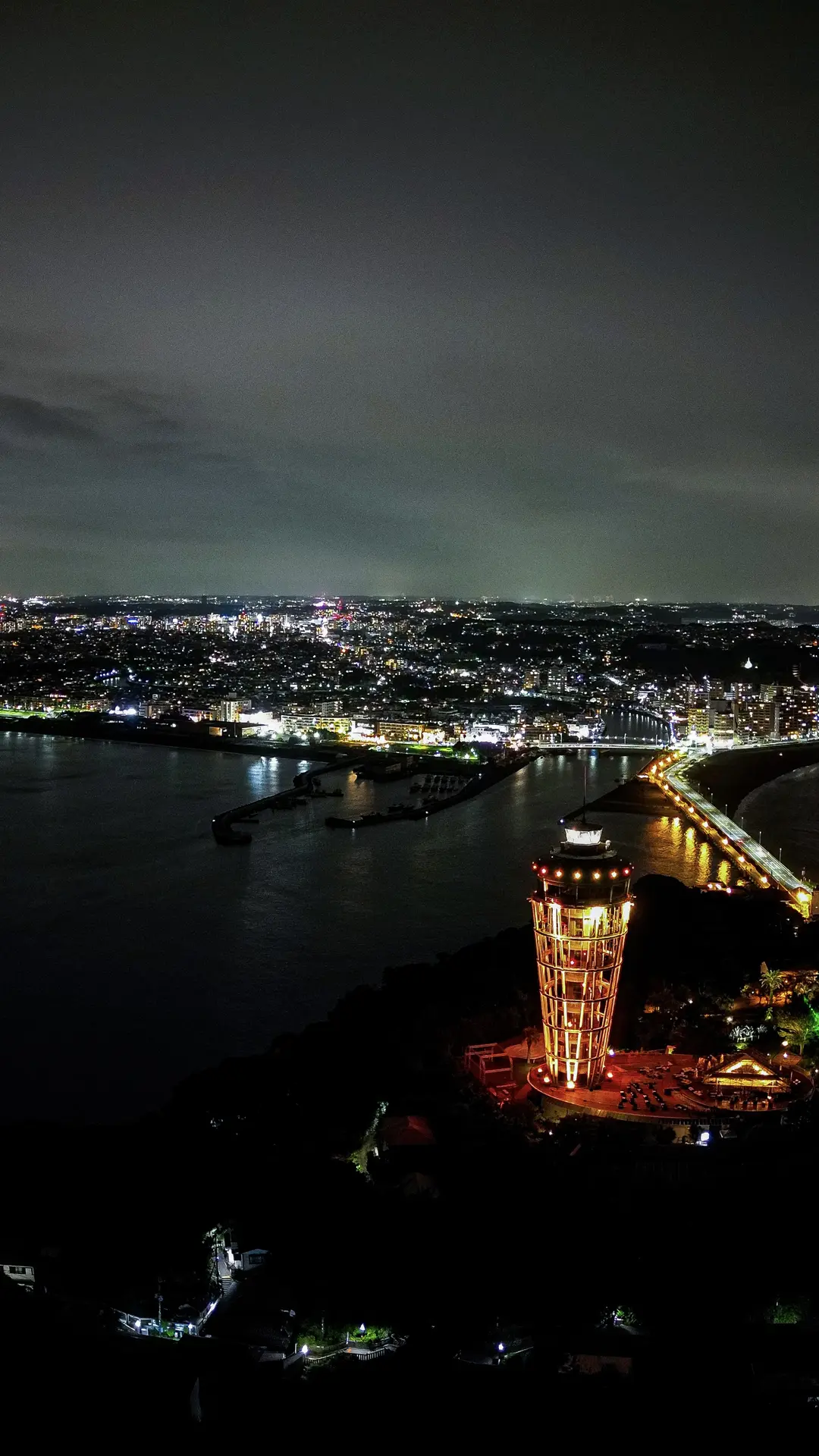 Aerial night view of the Enoshima Sea Candle lighthouse tower lit orange against the dark island, with the Pacific bay and city lights of Fujisawa stretching to the horizon