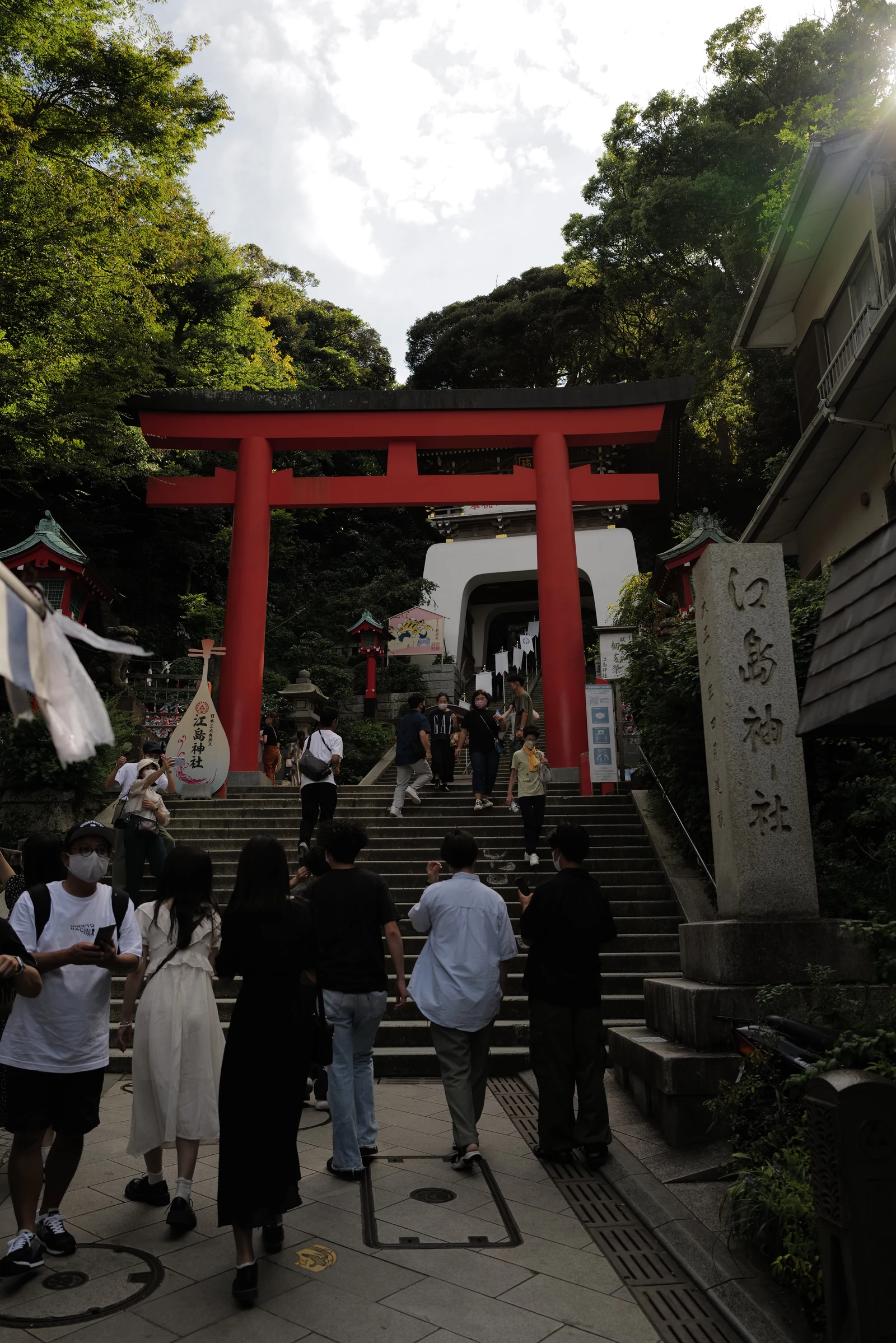 Red wooden torii gate of Enoshima Shrine at the bottom of a stone staircase, with visitors walking up the steps toward the gate, photographed in late afternoon golden light with the wooden sign reading 江島神社 (Enoshima Jinja) visible at right