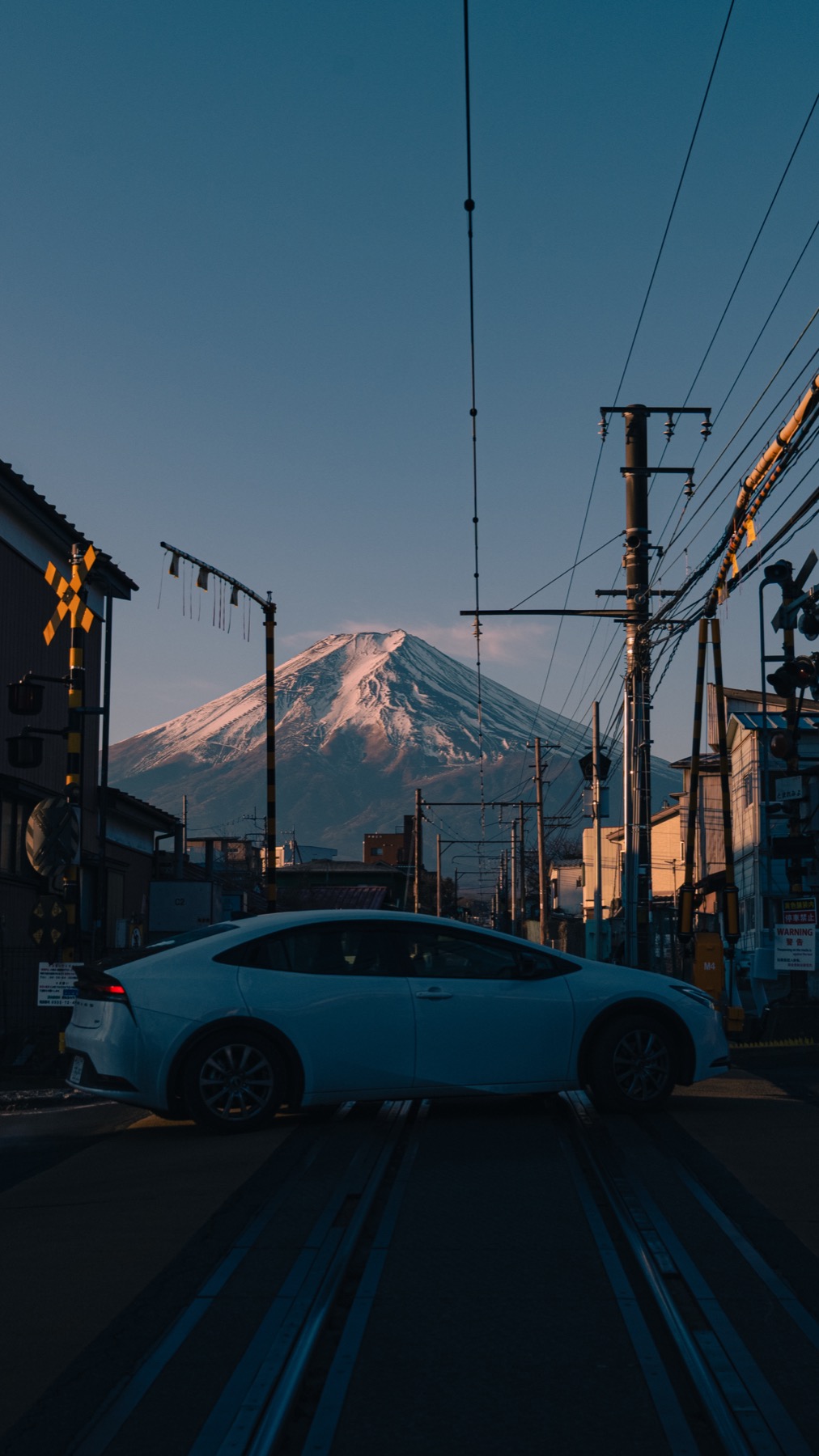 White Toyota Prius crossing a Fujikyu Railway level crossing at dawn with Mt Fuji visible behind glowing in pink red alpenglow, photographed February 6 2026 in Fujiyoshida