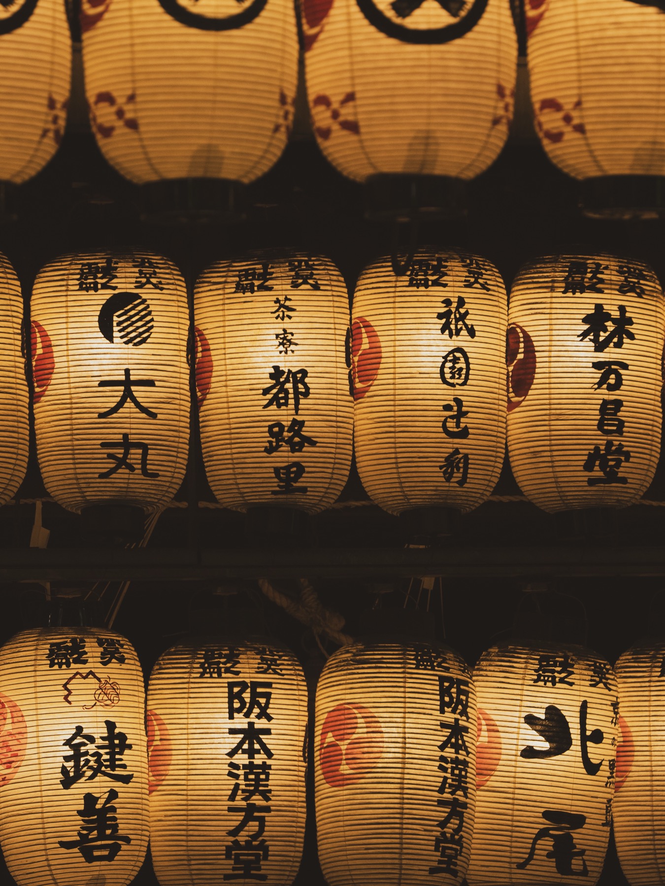 Close-up detail of dozens of cream coloured paper lanterns hanging in stacked rows each one hand brushed with the name of a neighborhood shop or sponsor in Japanese characters black ink on the cream paper face and a red crest at the base creating a wall of glowing patterned chochin during Gion Matsuri Yoiyama