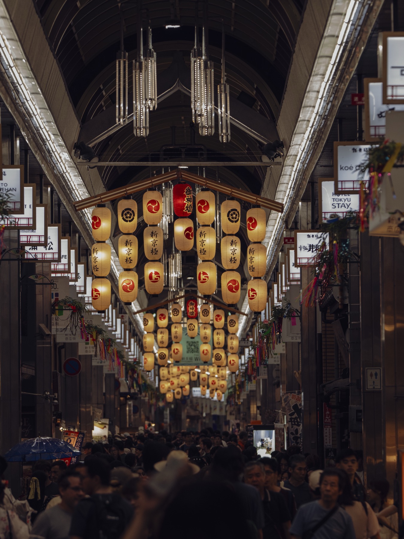 An interior view of the Shinkyogoku covered shopping arcade in Kyoto during Gion Matsuri with a long row of golden paper lanterns suspended from the ceiling each printed with kanji characters and red mitsudomoe crests, dense crowd of yukata-wearing visitors walking below