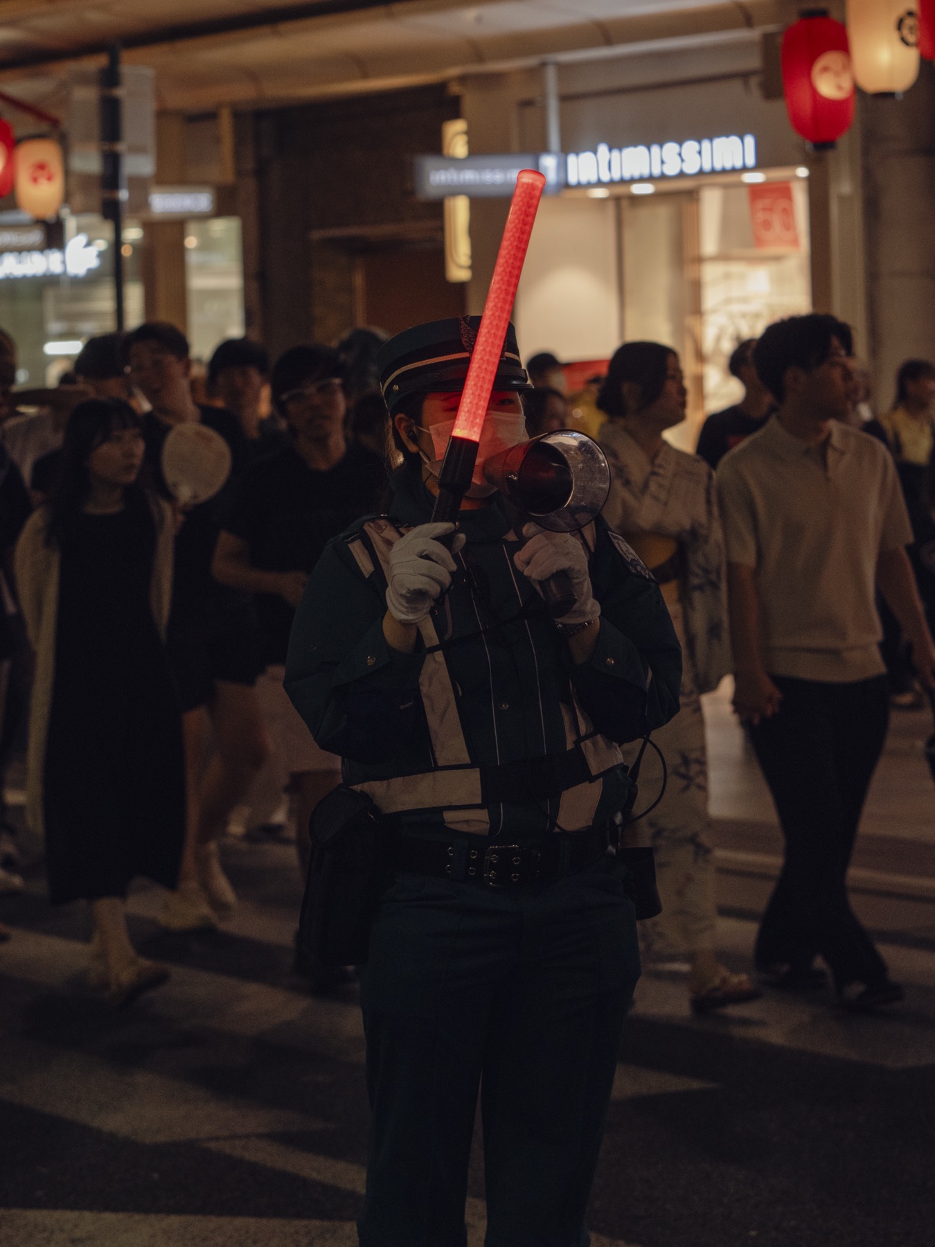 A uniformed Japanese police officer wearing a face mask and gloves stands in the middle of a Kyoto street during Gion Matsuri at night holding a red illuminated traffic baton and a megaphone directing the flow of yukata-wearing festival visitors past Intimissimi shop signs and red Japanese lanterns hanging overhead