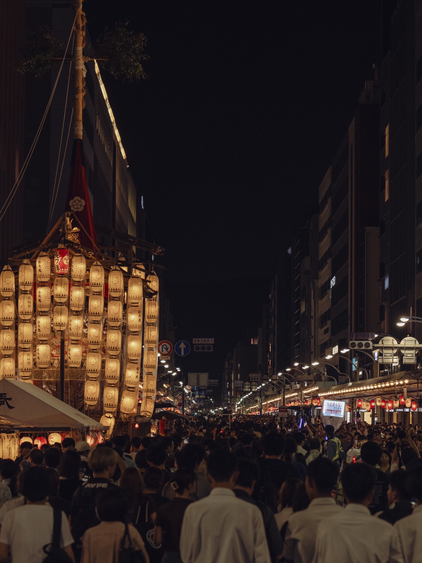 A tall vertical column of golden paper lanterns hanging from the upright pole of the Tsuki Hoko a yamaboko float on the streets of central Kyoto during Gion Matsuri Yoiyama with shop signs and the dense night-time festival crowd visible at the base