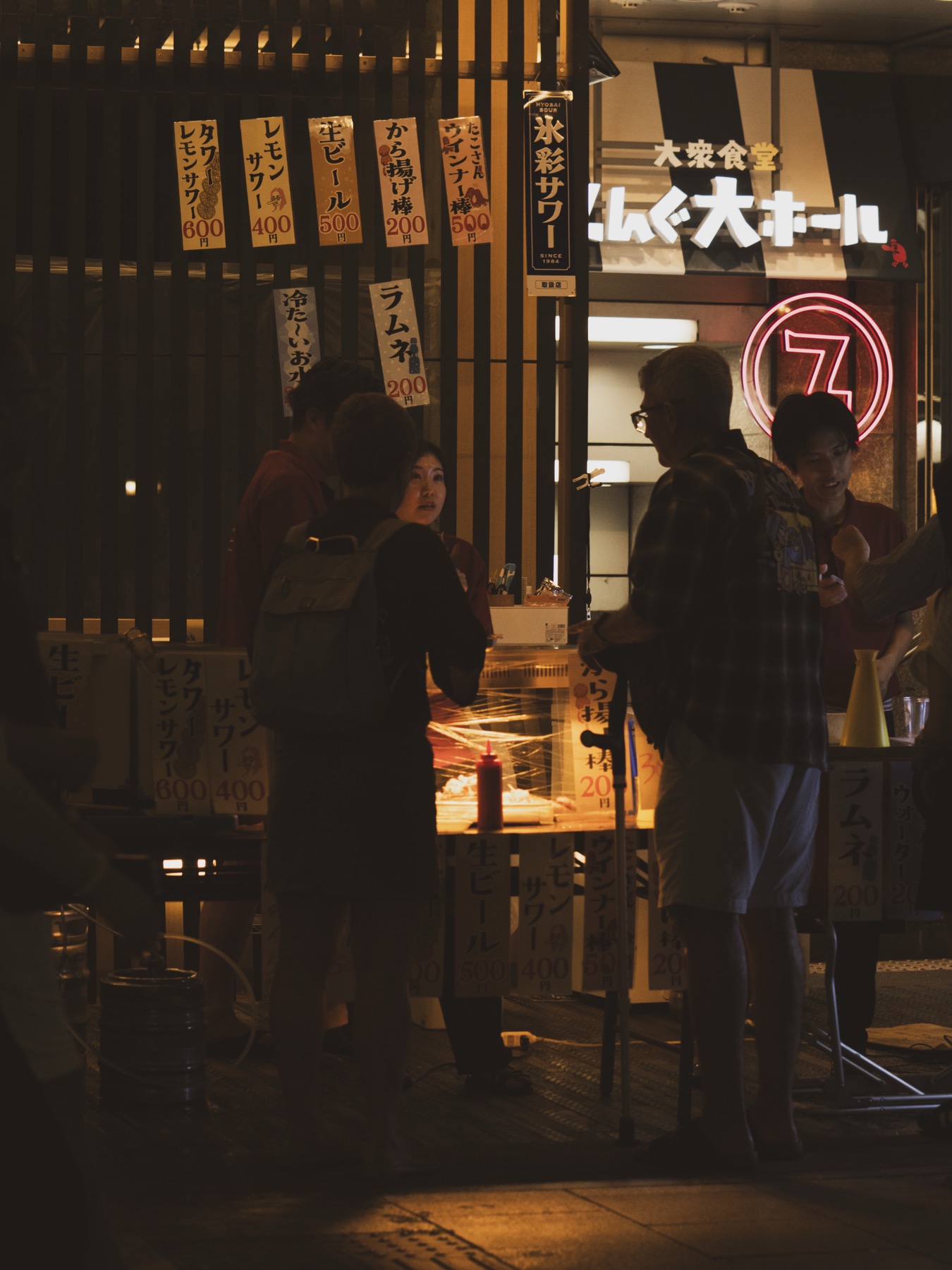 A small Gion Matsuri yatai food stall at night with handwritten paper price signs hanging on the front advertising lemon sour cocktails karaage chicken and yakitori in Japanese around 400 to 500 yen each with vendors and customers visible at the counter under warm overhead lights