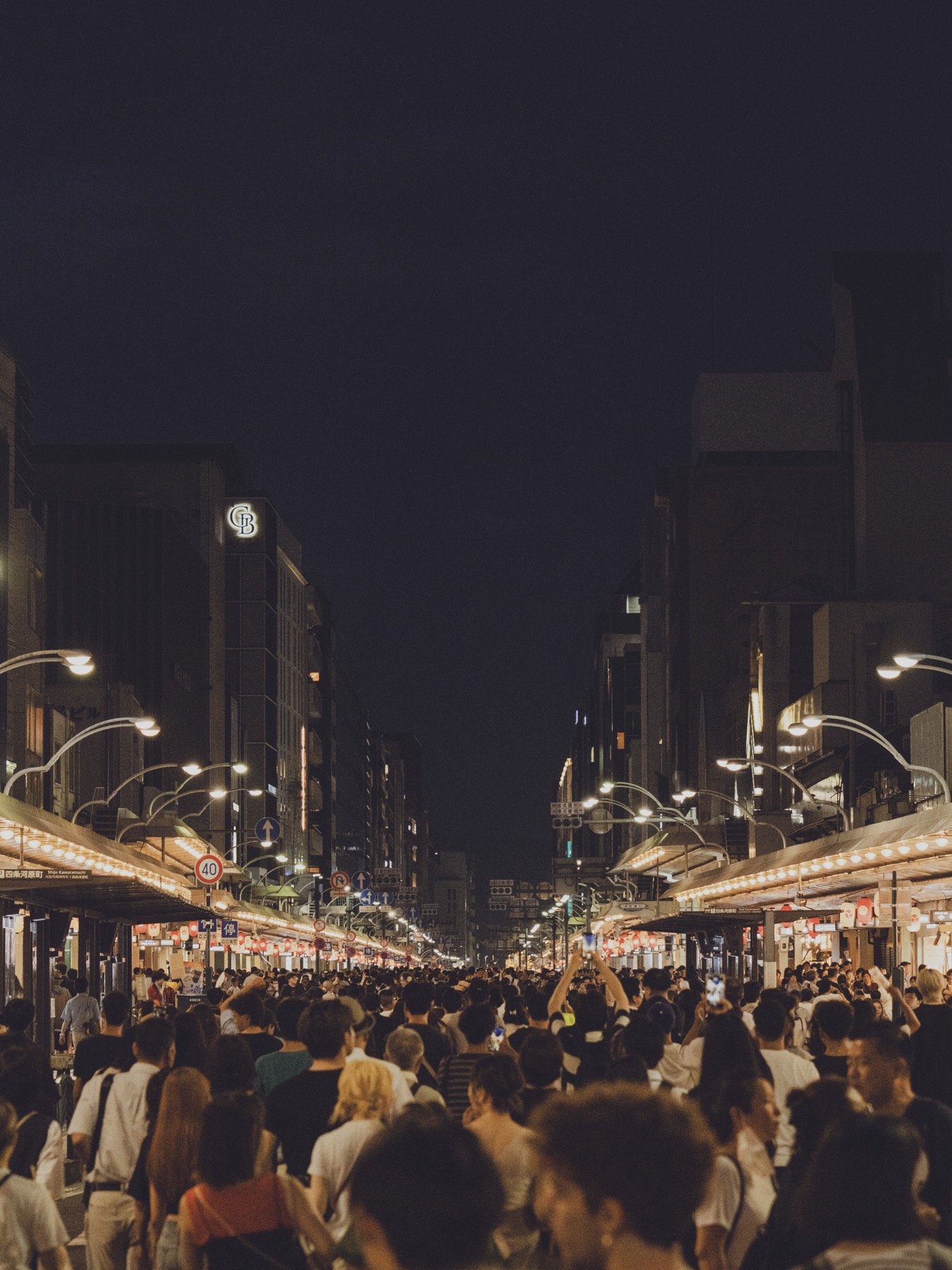 Wide panoramic view down Shijo-dori in Kyoto during Gion Matsuri Yoiyama night showing a dense crowd of thousands of festival visitors stretching down the closed street with rows of red and white yatai food stalls on both sides and tall buildings lit up overhead