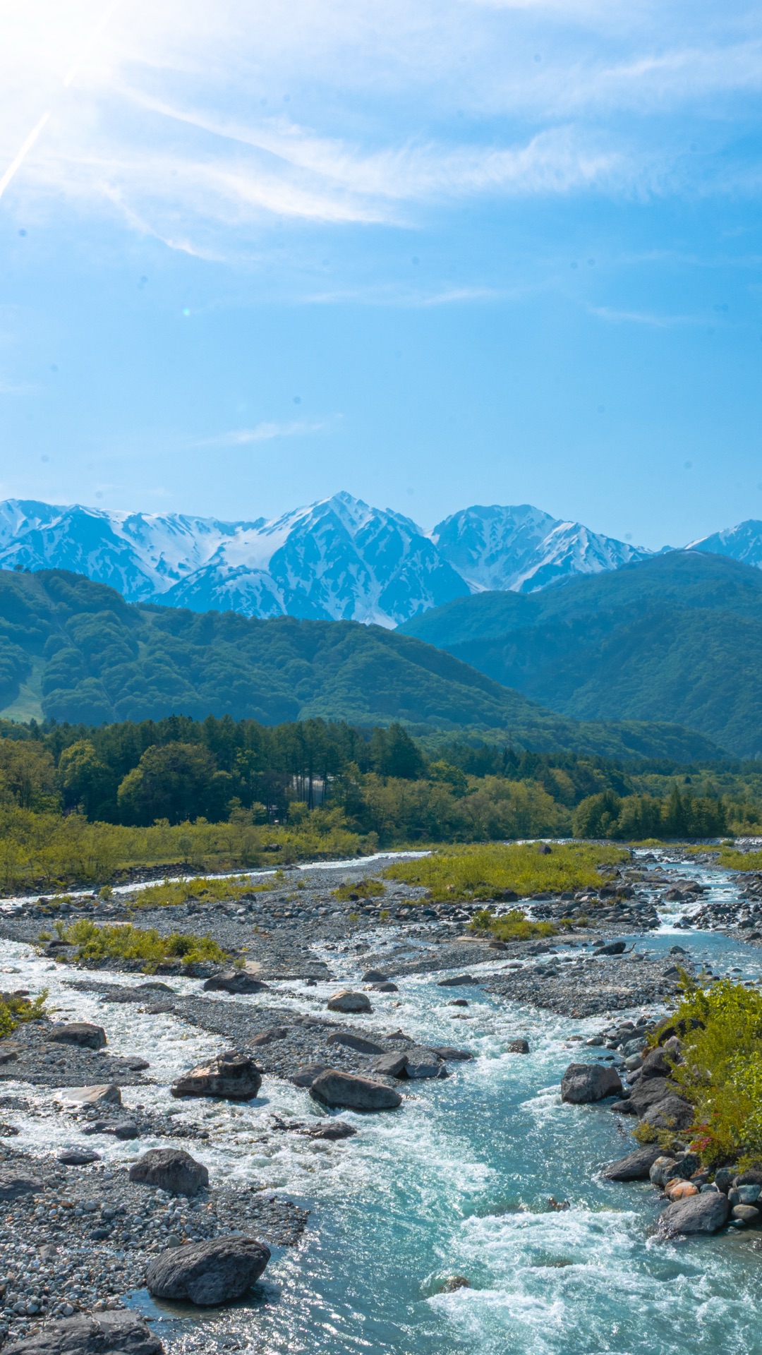Vertical composition of Hakuba Sanzan peaks with morning sun glare above and the Matsugawa river in the foreground