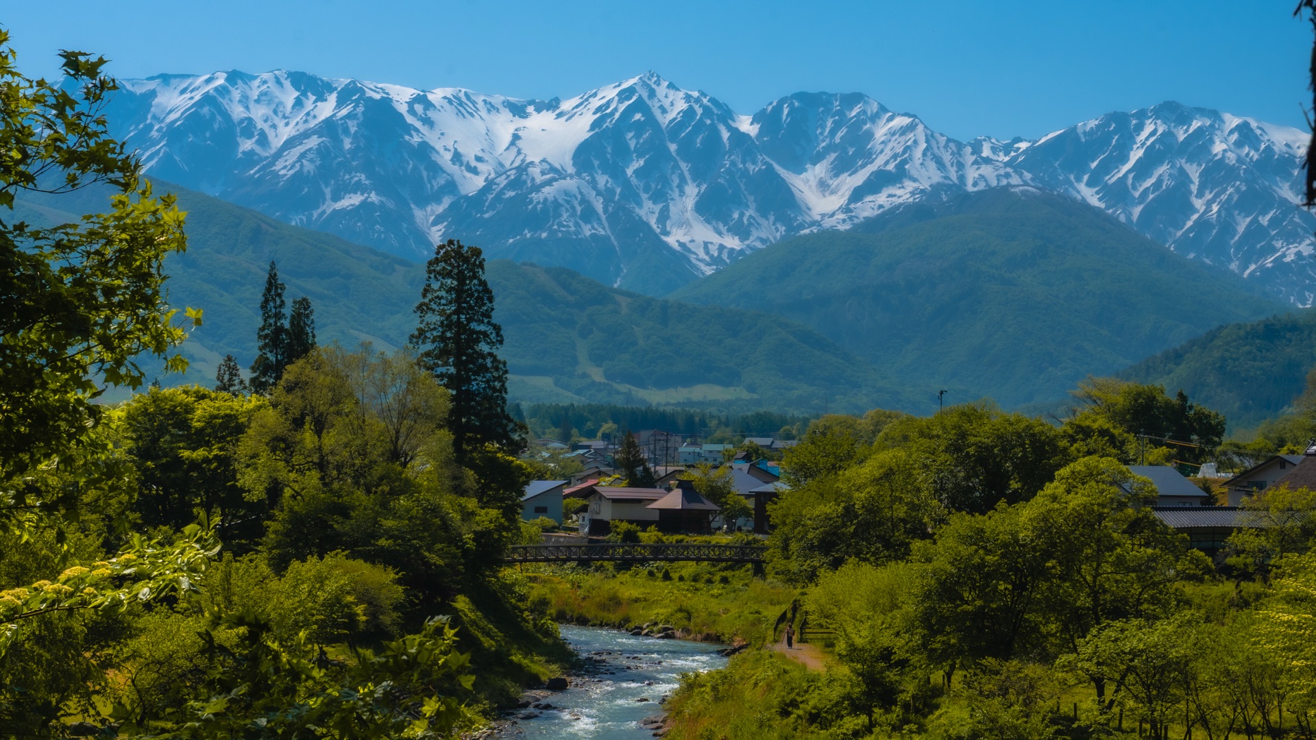 Wide view from Hakuba Oide Park showing snow-capped peaks, river, village rooftops and green forest