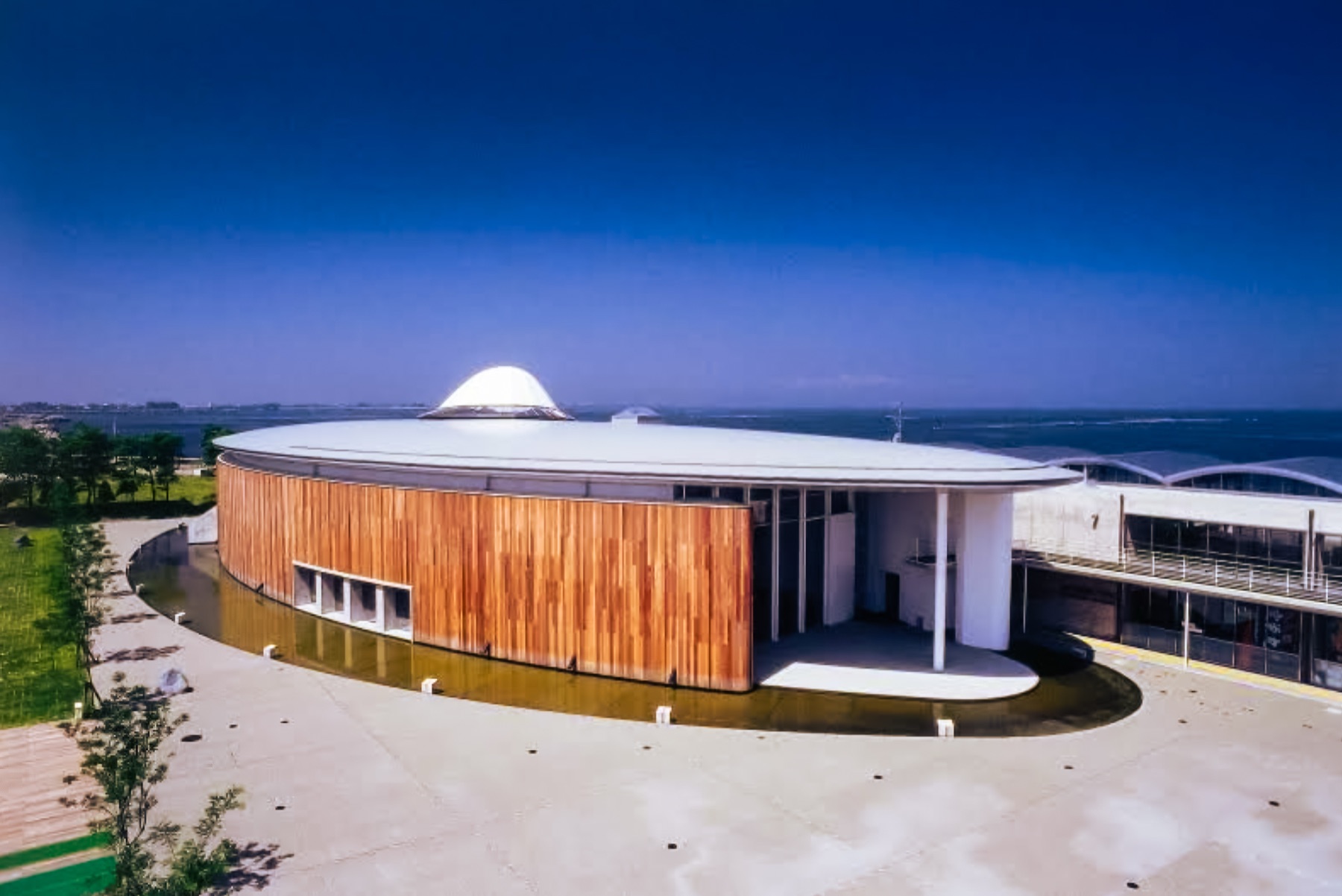 The circular wood-clad Hotaruika Museum building with a white domed roof, set beside the Sea of Japan under a clear blue sky at Wave Park Namerikawa in Toyama.