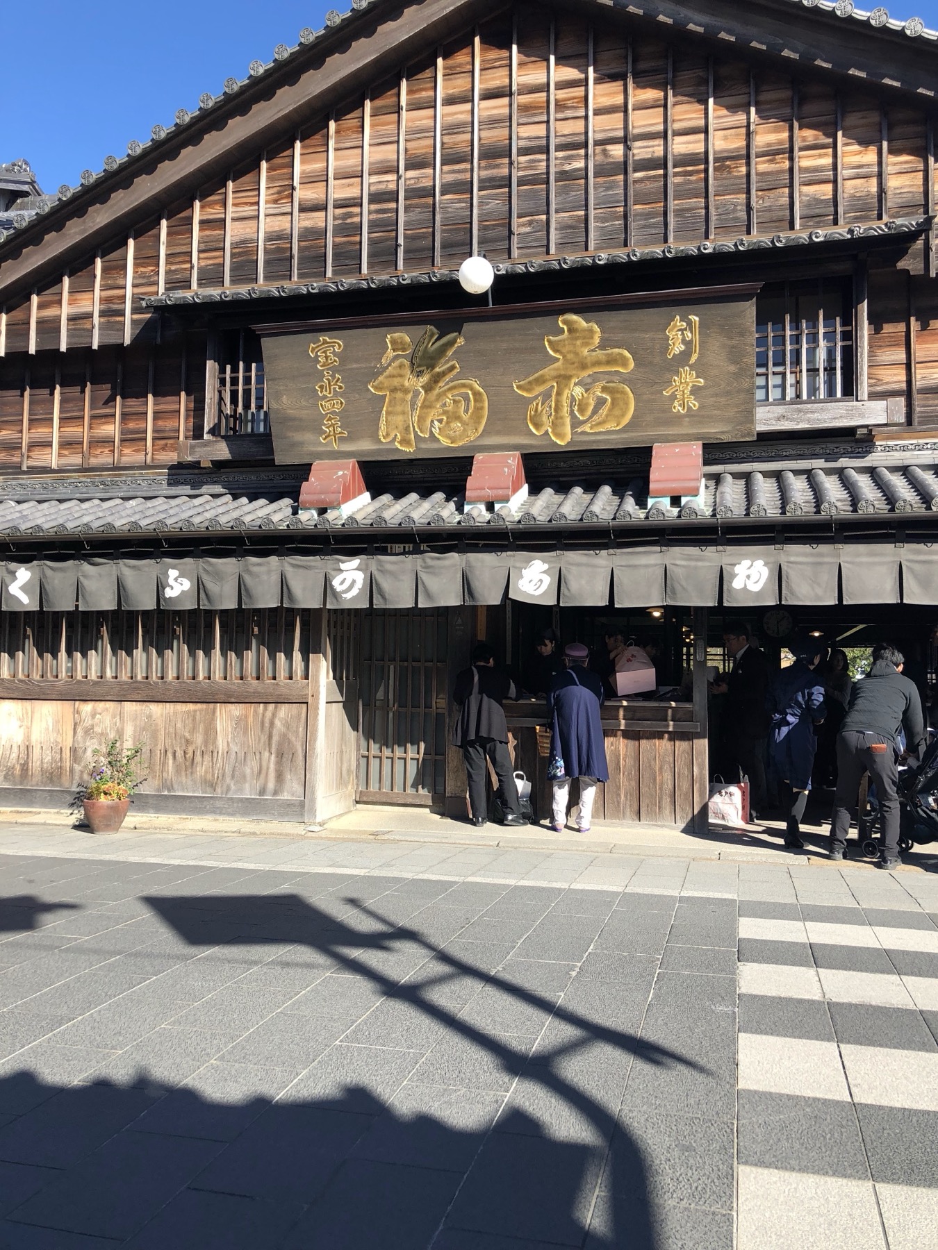 The wooden storefront of Akafuku Honten the original main store of the famous red bean rice cake shop on Oharaimachi street near Ise Jingu with the distinctive vermillion sign reading 赤福 in gold characters and customers entering through the noren curtained entrance