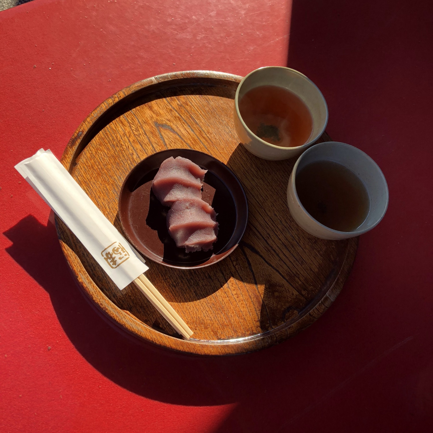 A round wooden tray with two small dishes of Akafuku red bean rice cake the signature smooth sweetened azuki paste over soft mochi served with two ceramic cups of hot green tea and chopsticks on a red lacquered counter at the Akafuku Honten main store in Ise