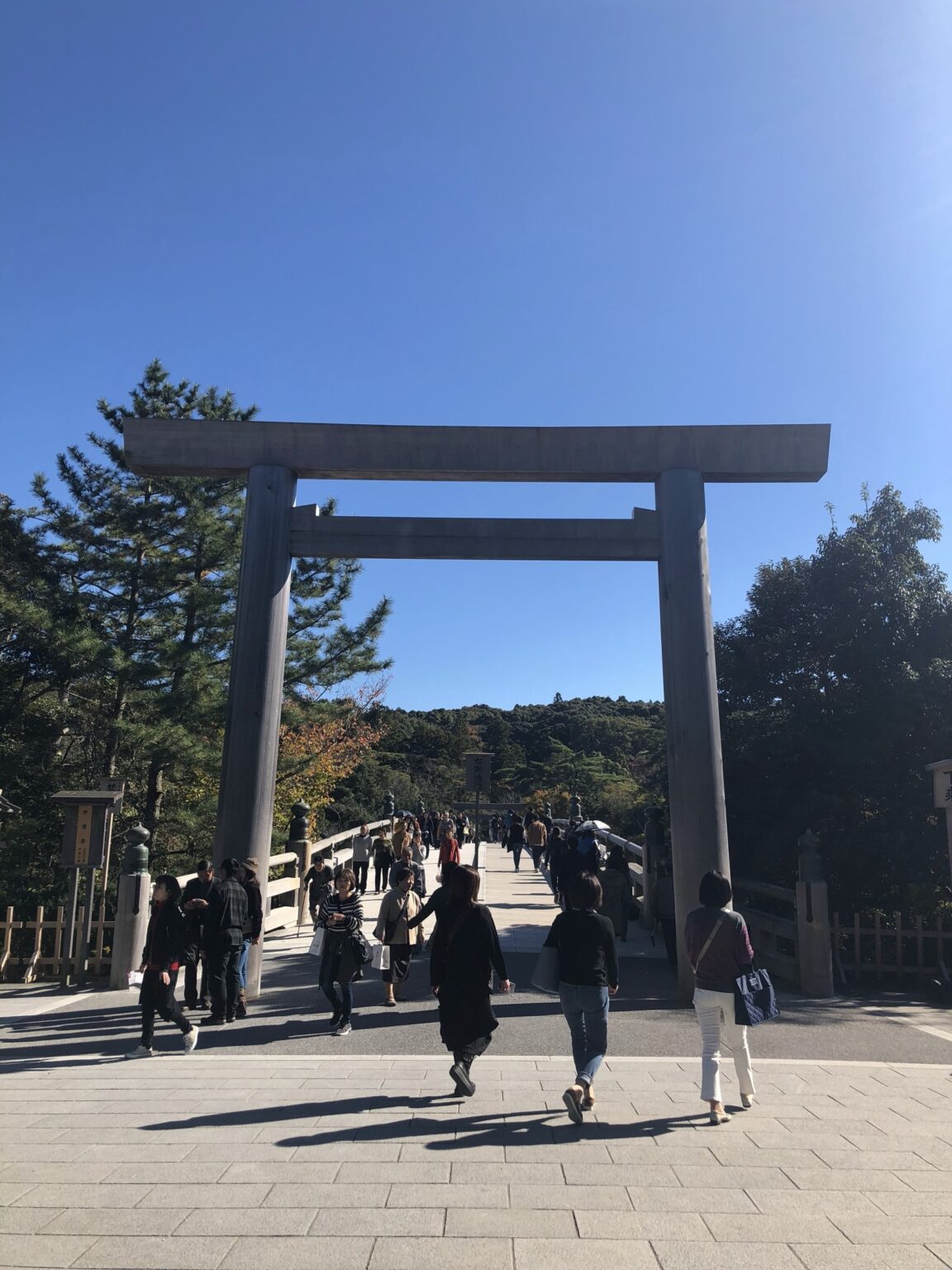 Massive wooden torii gate at the entrance of Ise Jingu Naiku with visitors crossing the Uji Bridge under a clear blue late-autumn sky, the sacred crossing point into Japan's most important Shinto shrine, low afternoon sun raking across the bridge planks