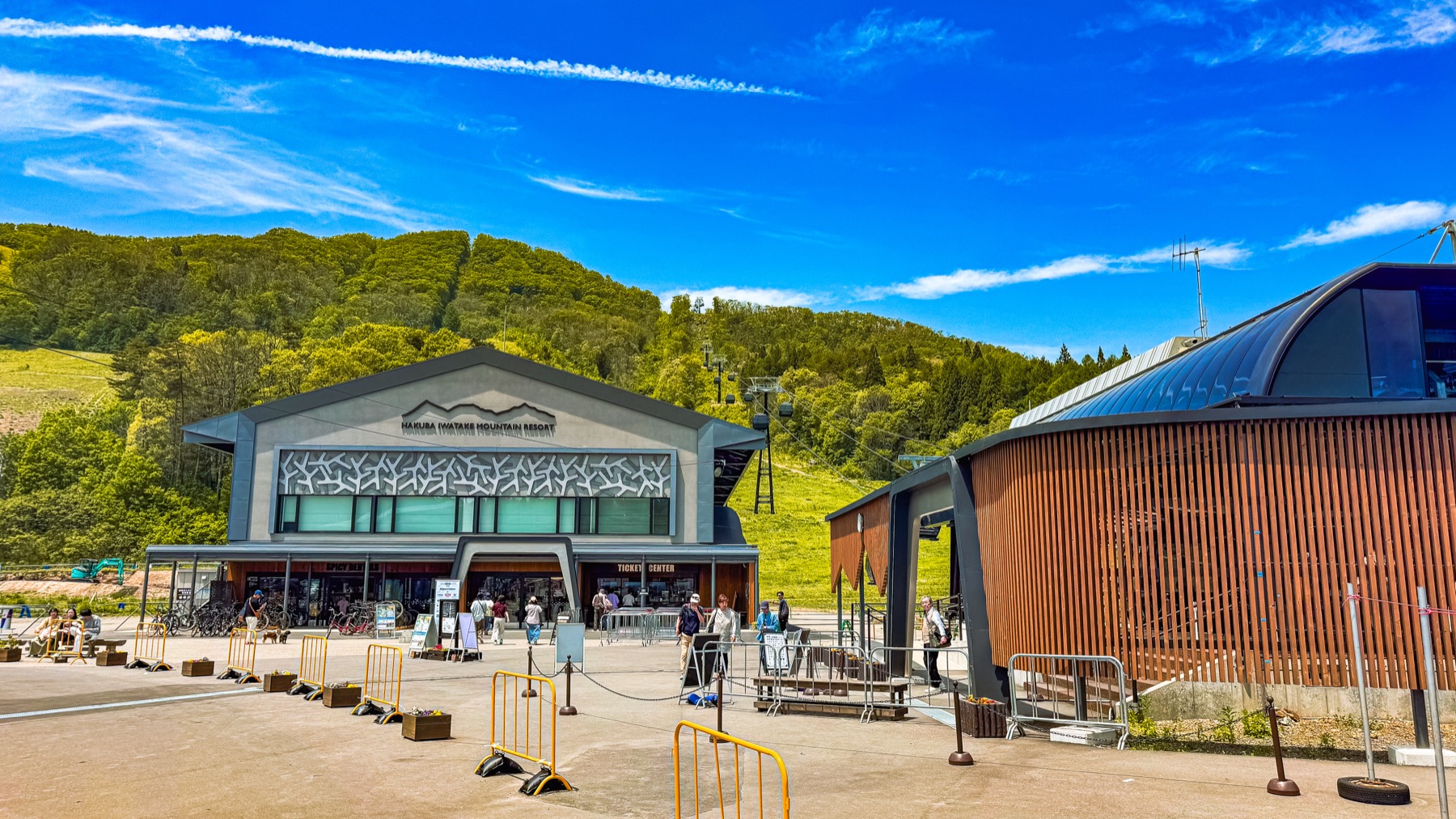 The exterior of the Hakuba Iwatake Mountain Resort gondola base station building under a bright blue sky with white clouds visitors gathered at the entrance with the green ski slope and forest visible behind the timber lodge style modern building at 750 metres elevation