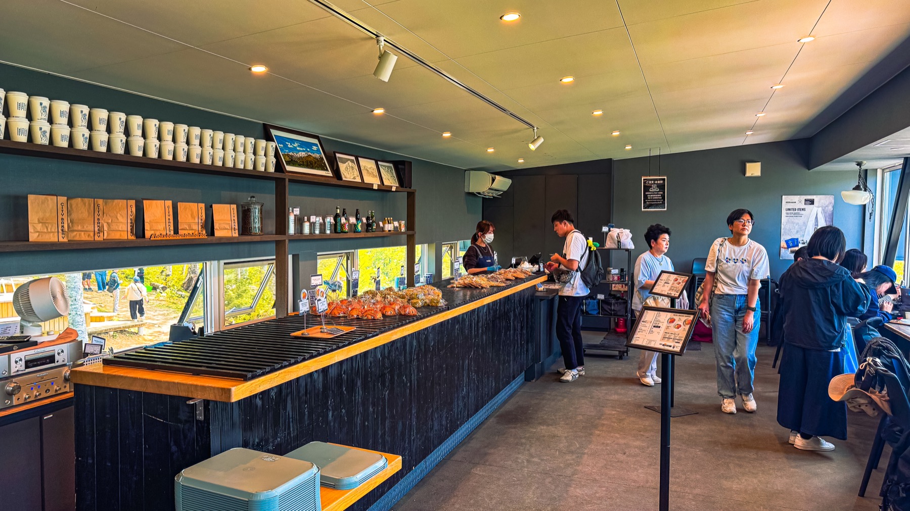 The interior of The City Bakery at Hakuba Mountain Harbor showing the wooden display counter stacked with fresh croissants and biscuits a row of takeaway coffee cups on shelves and several customers ordering at the counter with the menu visible on the wall above