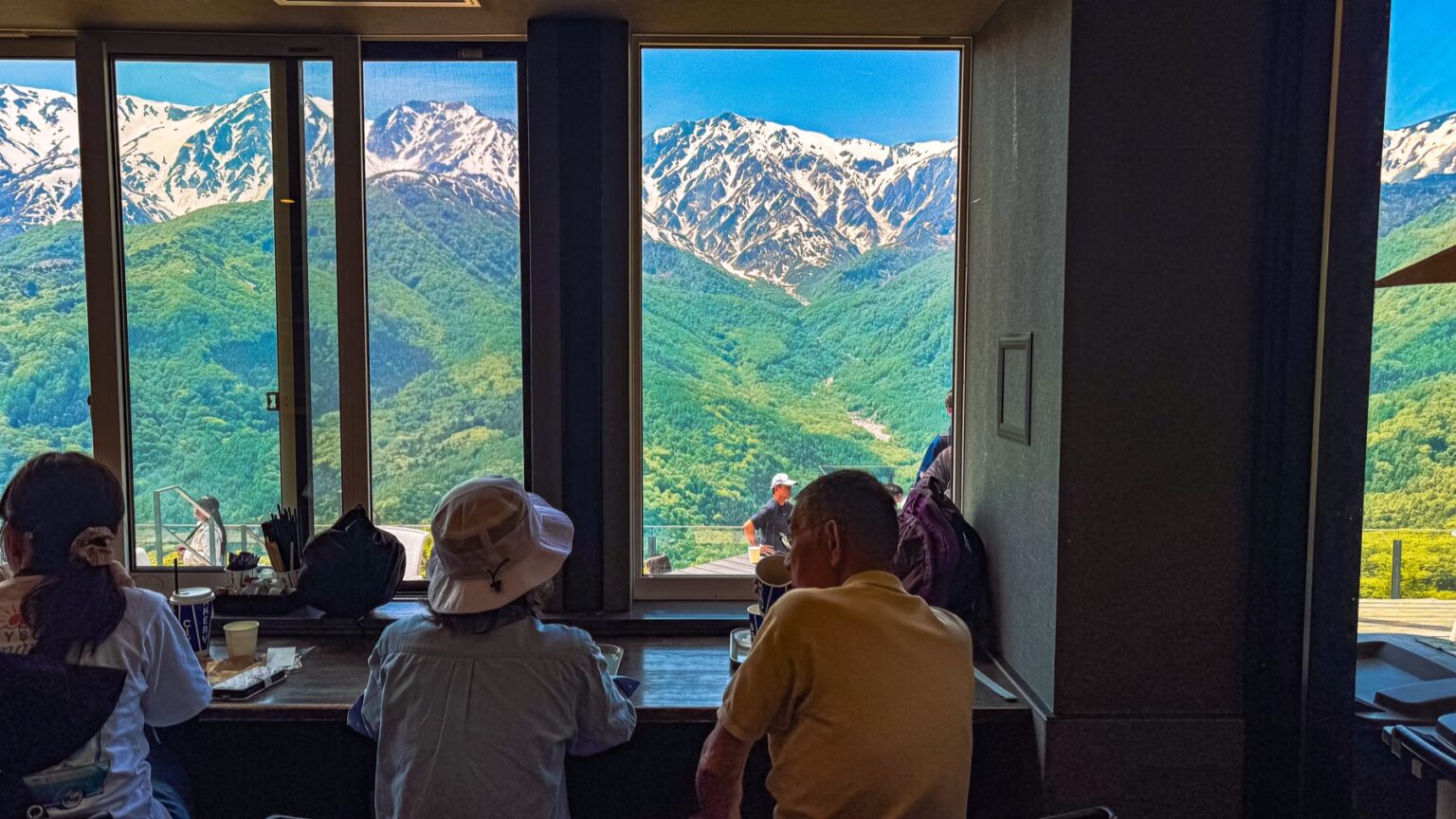 Indoor counter seating at The City Bakery Hakuba Mountain Harbor with several customers sitting at a wooden bar facing the floor to ceiling window that frames a wide view of the snow capped Hakuba Sanzan three peaks directly across the deep green valley