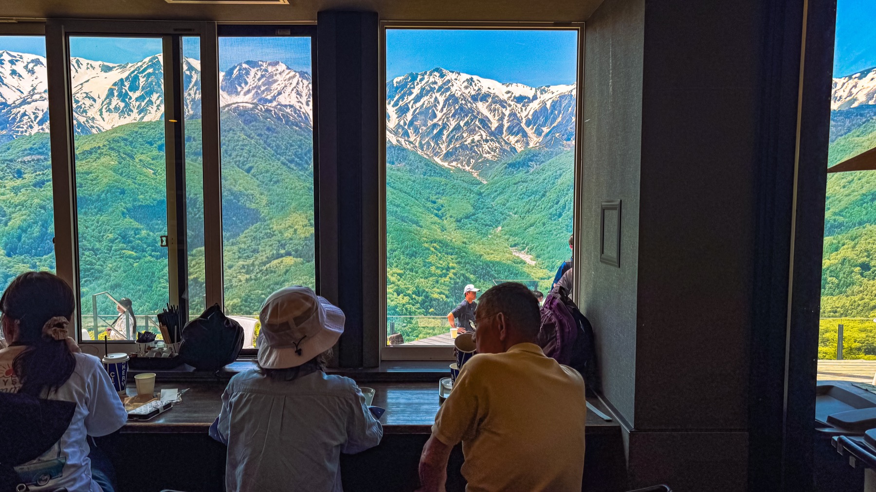 Indoor counter seating at The City Bakery Hakuba Mountain Harbor with several customers sitting at a wooden bar facing the floor to ceiling window that frames a wide view of the snow capped Hakuba Sanzan three peaks directly across the deep green valley