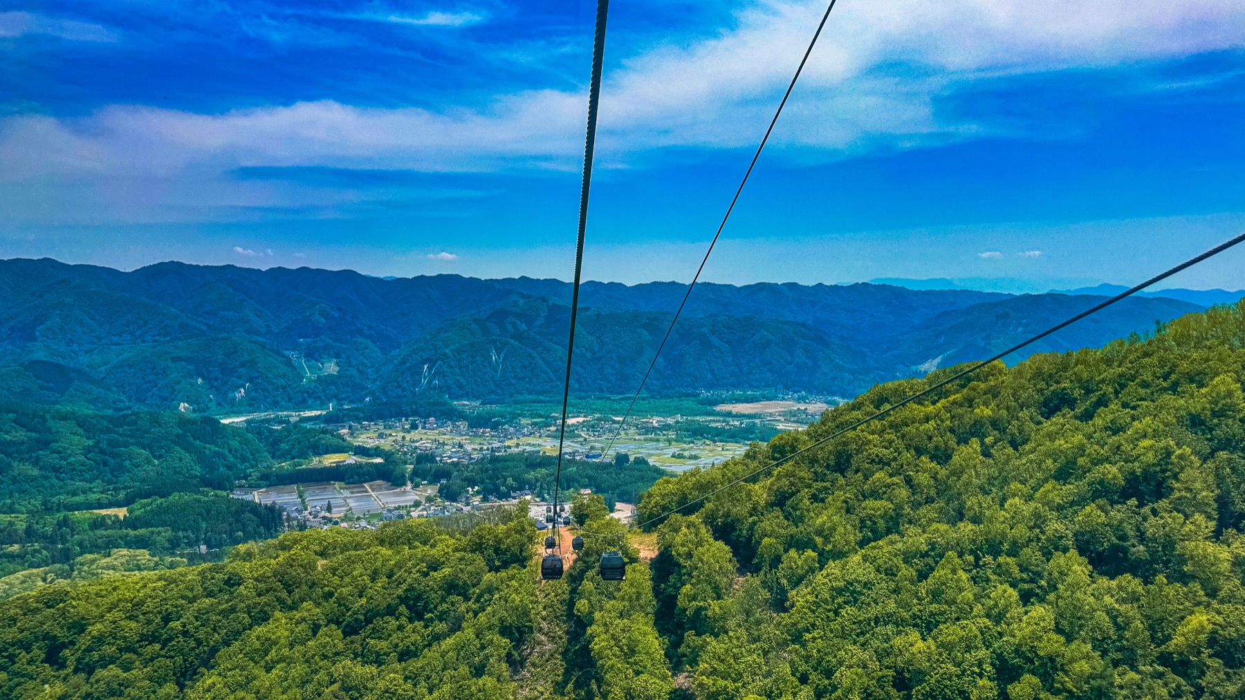 An aerial view from the Iwatake gondola Noah looking down on the entire Hakuba village basin with the cable suspension visible the village rooftops fish ponds and rice paddies far below and the distant Northern Alps stretching across the horizon