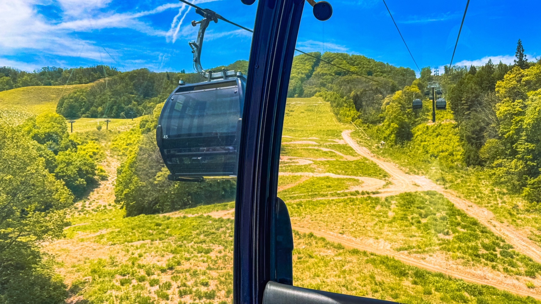 A view from inside the Noah gondola at Hakuba Iwatake Mountain Resort showing another black gondola passing in the opposite direction above the bright green summer ski slope with cable system and support tower visible on the way up to the 1289 metre summit