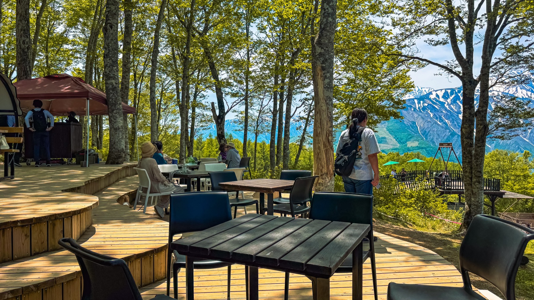 Hakuba Mountain Harbor outdoor cafe deck with wooden tables and folding chairs scattered under tall beech trees a red parasol providing shade and visitors browsing the view of the Hakuba Sanzan peaks visible through the foliage on the right