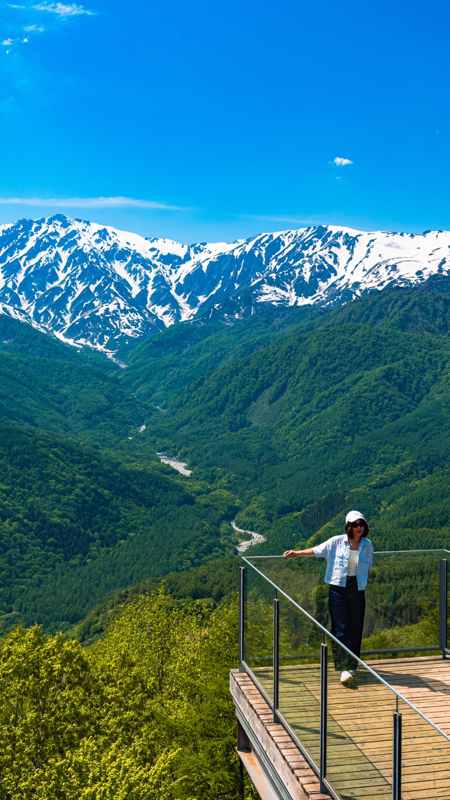 A visitor in white standing at the end of the wooden cantilevered Yahho Deck at Hakuba Iwatake Mountain Resort one arm extended pointing toward the snow capped Hakuba Sanzan three peaks across the deep green valley the signature shouting yahho spot vertical composition