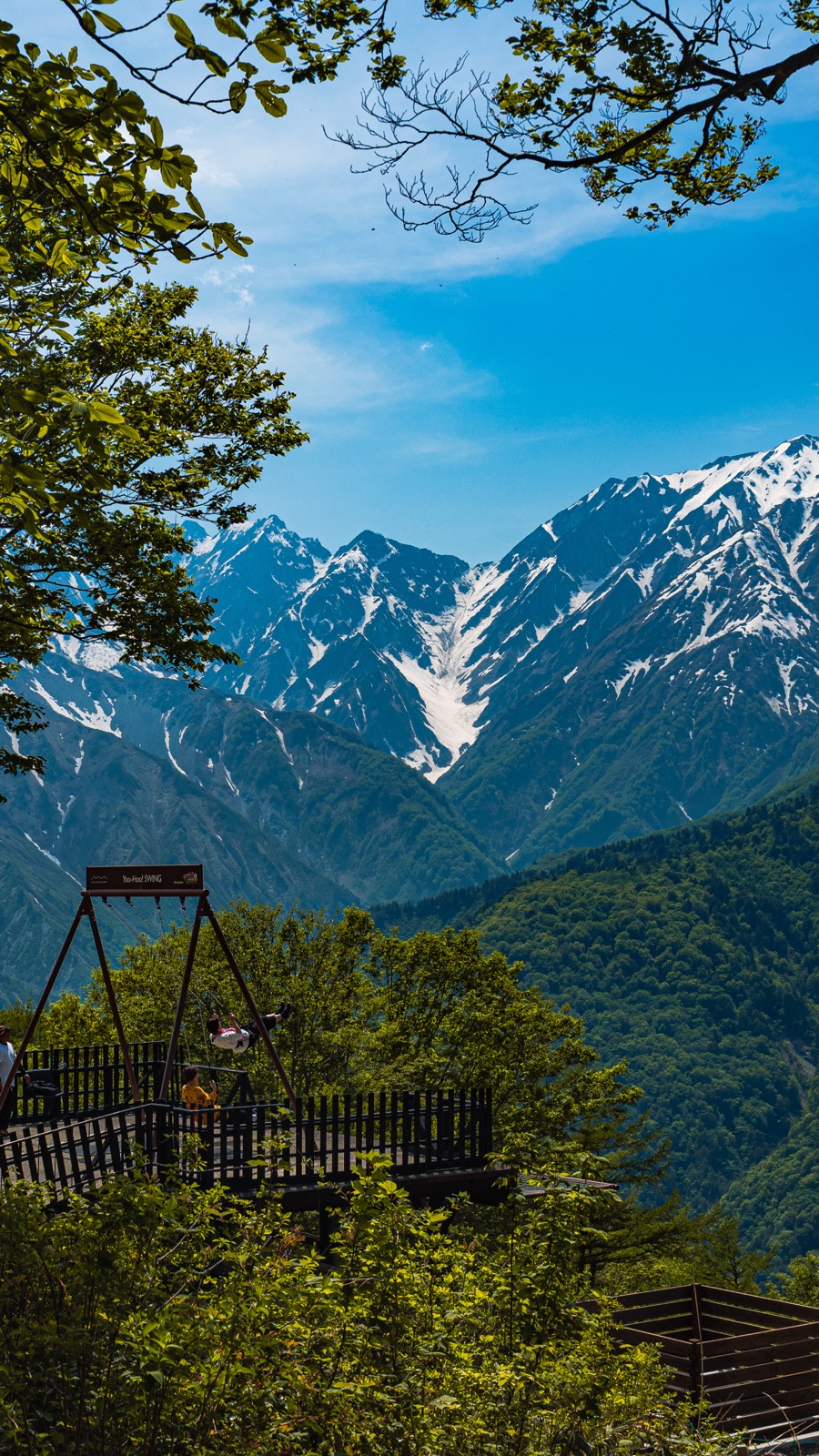 Vertical photograph of the Yahho Deck cantilevered wooden viewing platform at Hakuba Iwatake Mountain Resort with the snow-capped Hakuba Sanzan three peaks rising above green forest framed by beech tree branches in the upper foreground
