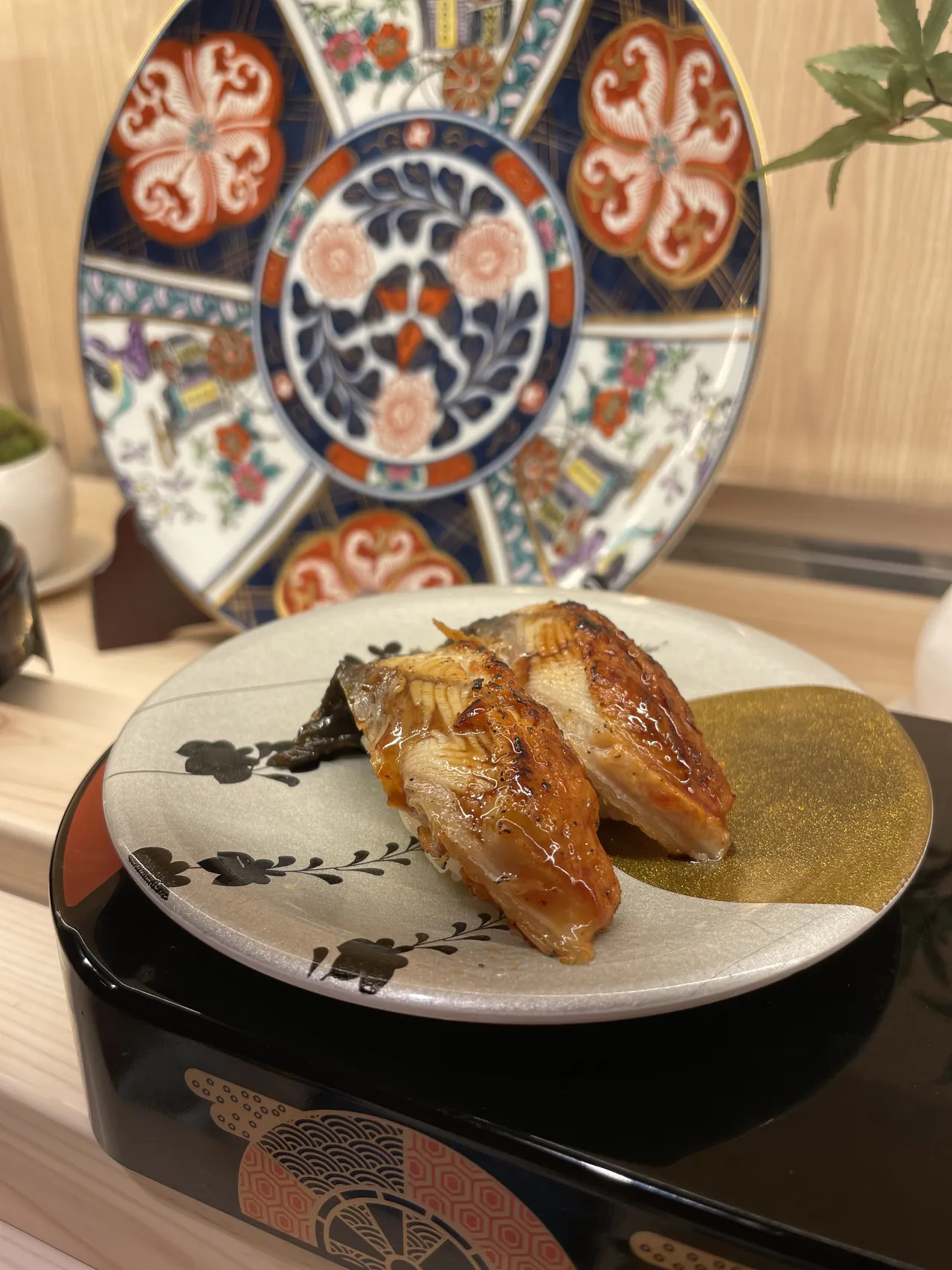 Two pieces of steamed anago conger eel glazed with house sauce, served on a black plate beside a green tea biscuit, with Imari porcelain in the background