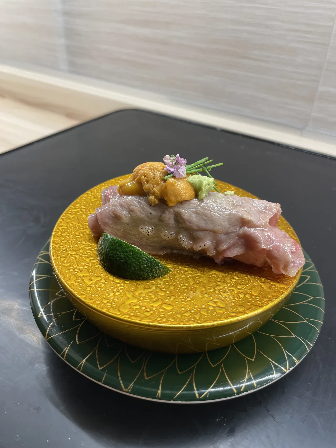 Tuna otoro draped over a bed of uni sea urchin on a gold and green ceramic plate, with sudachi citrus wedge and wasabi