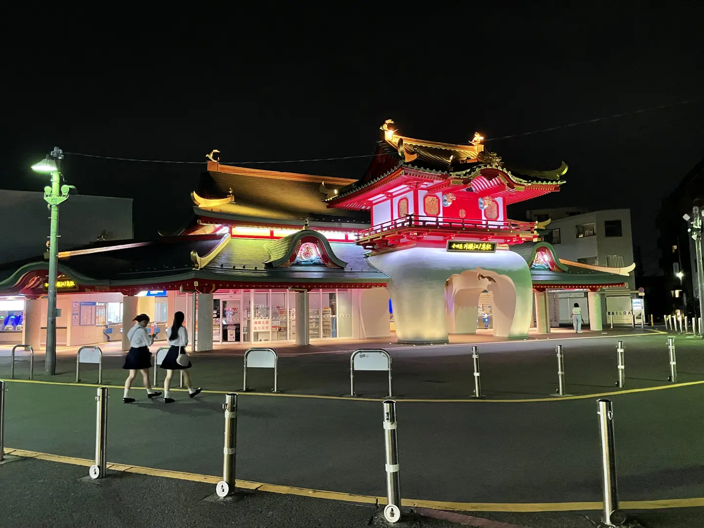 Katase-Enoshima Station at night — a recreation of the Dragon Palace from Japanese folklore in red, white and gold, illuminated against the dark night sky, with two schoolgirls in uniform walking past in the foreground