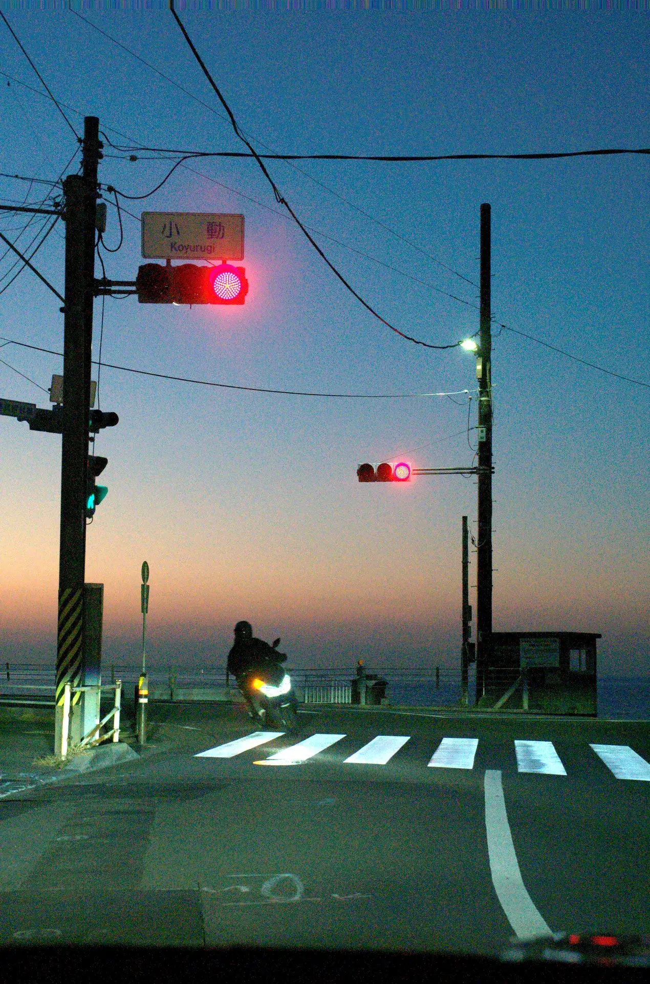 The Koyurugi traffic light intersection in Shonan at predawn with a motorcyclist crossing and the Pacific Ocean visible behind