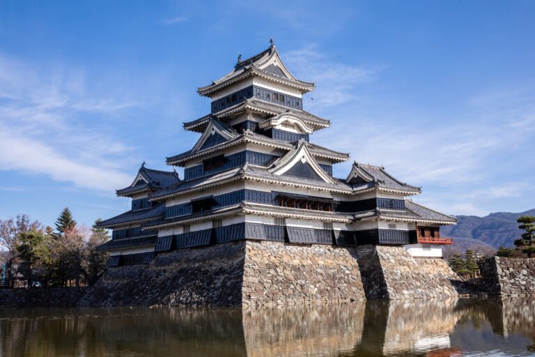 The black five-storey keep of Matsumoto Castle in Nagano reflected in the still water of its inner moat under a clear sky, with the stone base rising straight from the water and bare early-spring trees along the far bank.