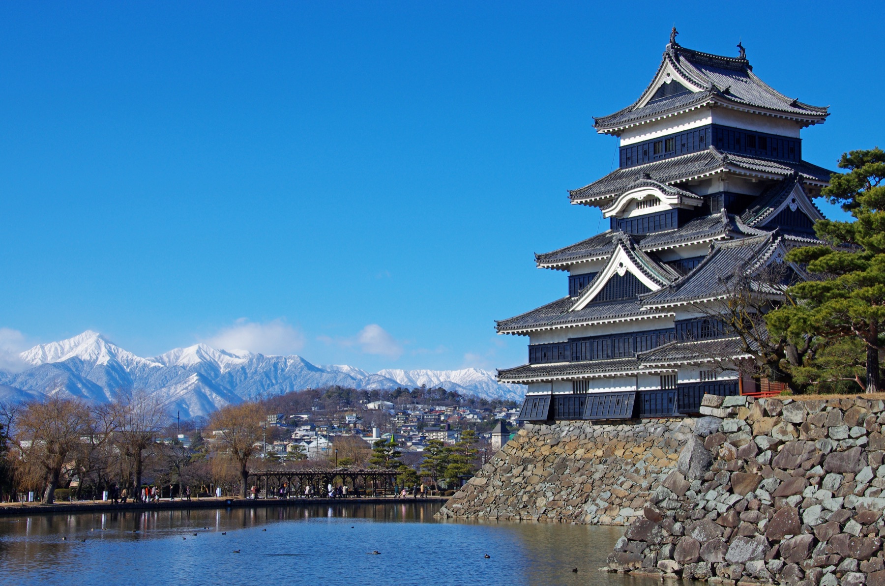 Matsumoto Castle black keep on the right with the snow-covered peaks of the Northern Japan Alps rising behind the town across the moat on a clear winter day, with visitors on the lakeside path.